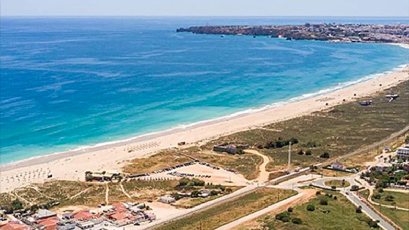 Acquista campeggi in Portogallo Vista aerea di una lunga spiaggia sabbiosa con onde turchesi, delimitata da aree erbose e strade, con alcuni edifici, parcheggi e la possibilità di acquistare una casa vacanze o un campeggio vicino alla riva sotto un cielo limpido. 0121Management - l'agente immobiliare per campeggi e case vacanze