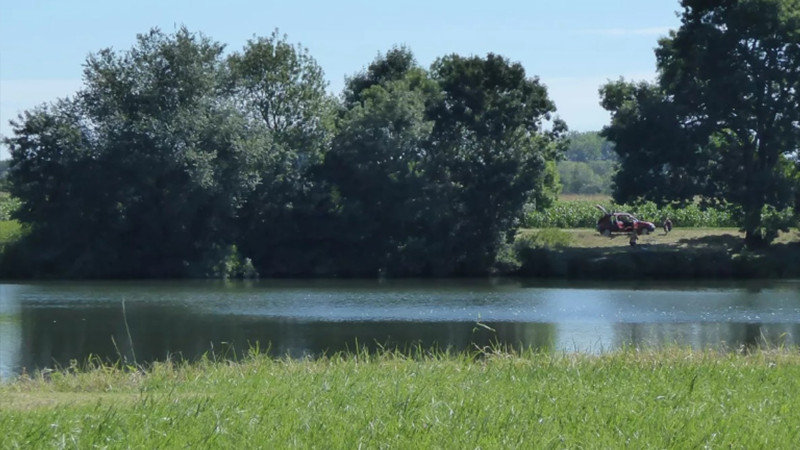 Un fiume tranquillo, costeggiato da erba verde e alberi alti, con una persona seduta accanto a una bicicletta rossa in una giornata di sole: l'ispirazione perfetta per la tua prossima casa vacanza o soggiorno glamping. 0121Management: l'agente di campeggi e case vacanza 