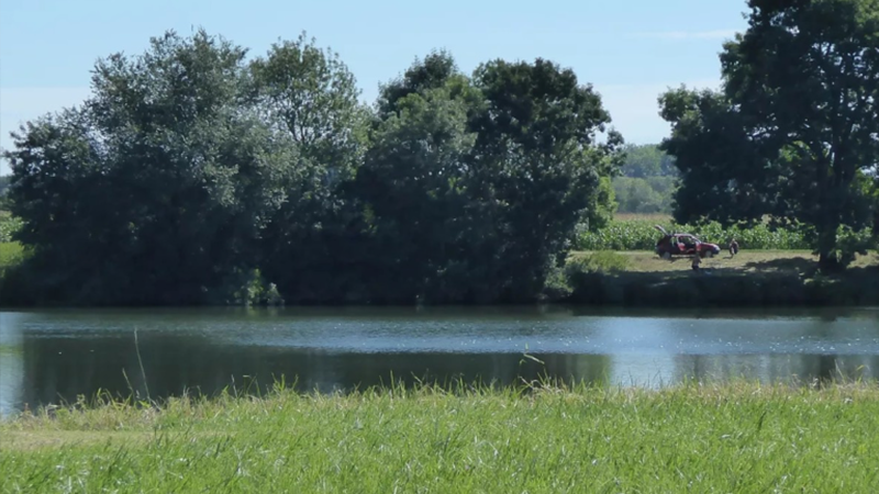 Ein ruhiger Fluss, gesäumt von grünem Gras und hohen Bäumen, mit einer Person, die an einem sonnigen Tag neben einem roten Fahrrad sitzt - die perfekte Inspiration für Ihre nächste Ferienimmobilie oder Ihren Glamping-Aufenthalt. 0121Management - der Campingplatz und Ferienimmobilien Makler
