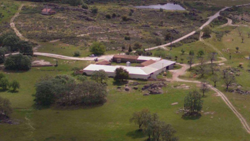 Acquista un campeggio in Portogallo Vista aerea di un complesso di edifici rurali con tetti bianchi, circondato da una vegetazione lussureggiante, alberi sparsi, tortuose stradine di campagna, terreno roccioso e un piccolo laghetto: perfetto come casa per le vacanze o da convertire in un esclusivo rifugio glamping. 0121Management: l'agente immobiliare per campeggi e vacanze