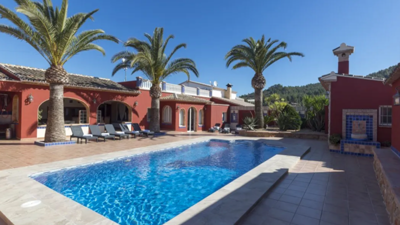 Eine große rote Villa mit Bögen und Palmen umgibt einen rechteckigen Swimmingpool unter einem klaren blauen Himmel. Ideal als Ferienimmobilie, lädt dieses Anwesen zum Investieren und Entspannen auf Sonnenliegen ein, die am Pool auf der gefliesten Terrasse aufgestellt sind. 0121Management - der Campingplatz und Ferienimmobilien Makler