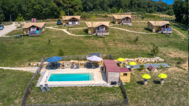 Vista aerea di un campeggio glamping rurale con sei tende di lusso su una collina erbosa e una piscina rettangolare recintata con lettini e ombrelloni in primo piano: una posizione perfetta per chi desidera investire o acquistare un campeggio. Sono visibili alberi e auto parcheggiate. 0121Management – ​​l'agenzia di intermediazione per campeggi e immobili per vacanze.  