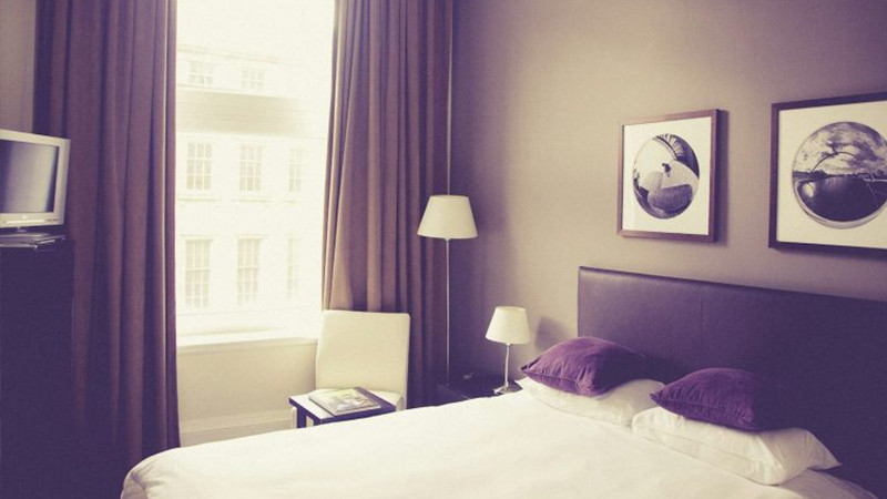 Acquista catena alberghiera in Germania Un'accogliente camera d'albergo con un letto ben fatto, due cuscini lilla, una lampada da comodino e un arredamento ispirato al glamping offre un livello di comfort che ricorda un campeggio di lusso. Rilassatevi vicino all'ampia finestra con le sue tende marroni e godetevi lo spazio progettato con cura. 0121Management - l'agenzia immobiliare per campeggi e vacanze