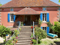 Eine charmante rosa Ferienimmobilie mit rotem Ziegeldach, blauen Fensterläden und einer Holztreppe, die zur Tür führt, umgeben von üppigem Grün und Topfpflanzen - perfekt, um in Ihren Traumurlaub zu investieren. 0121Management - der Campingplatz und Ferienimmobilien Makler