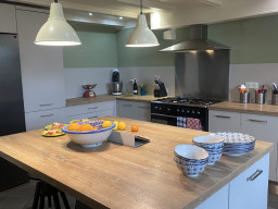 Eine moderne Küche mit einer hölzernen Inselarbeitsplatte, einer Obstschale, gestreiften und gemusterten Schalen bietet Luxus im Glamping-Stil. Hängelampen erhellen den Raum, während ein Herd mit Dunstabzugshaube diese einladende Ferienimmobilie vervollständigt. 0121Management - der Campingplatz und Ferienimmobilien Makler