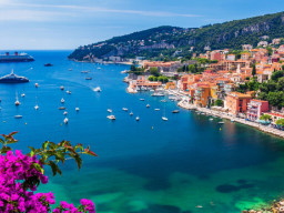 Eine lebhafte Küstenstadt mit farbenfrohen Gebäuden säumt eine türkisfarbene Bucht voller Boote und Yachten. Rosafarbene Blumen stehen im Vordergrund, während Sie auf den grünen Hügeln Campingplätze und ein Kreuzfahrtschiff unter dem klaren blauen Himmel entdecken können. 0121Management - der Campingplatz und Ferienimmobilien Makler