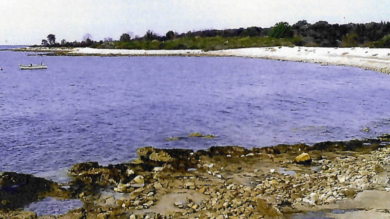 Acquista un campeggio in Croazia Una costa rocciosa con acque cristalline e azzurre che si incurva dolcemente, delimitata da una stretta spiaggia sabbiosa e da una vegetazione lussureggiante: un luogo ideale per investire in un campeggio o creare un rifugio glamping unico. Una piccola barca galleggia vicino al lato sinistro dell'immagine. 0121Management – l'agenzia di intermediazione per campeggi e immobili per vacanze