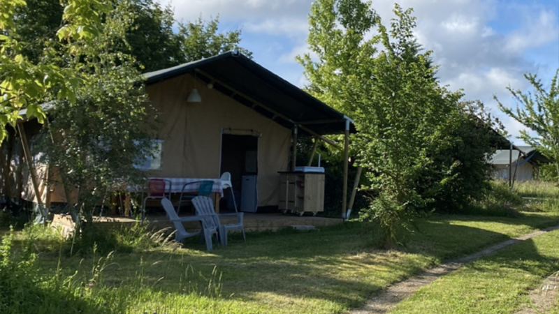 Ein Glamping-Zelt mit einer überdachten Veranda, Stühlen und einem Tisch steht zwischen grünen Bäumen und Gras unter einem teilweise bewölkten Himmel - die perfekte Inspiration für alle, die ihren eigenen Campingplatz kaufen oder verkaufen möchten. 0121Management - der Campingplatz und Ferienimmobilien Makler
