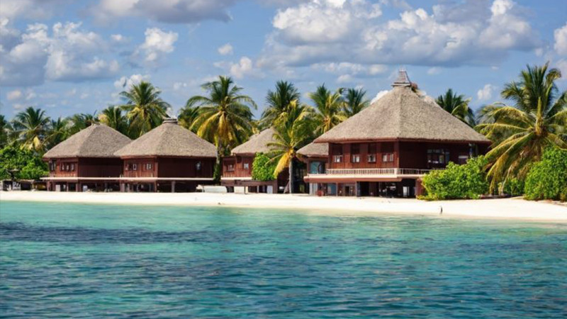 Acquista hotel alle Maldive Ville tropicali sulla spiaggia con tetti di paglia, circondate da palme, offrono un'esperienza unica di vacanza glamping con vista sulle acque turchesi e cristalline sotto un cielo parzialmente nuvoloso. 0121Management - l'agente immobiliare per campeggi e case vacanza