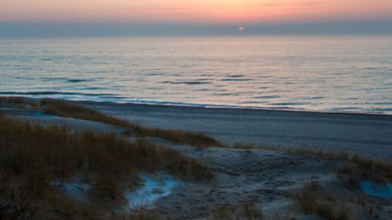 Sonnenuntergang über dem ruhigen Meer, mit Blick auf sandige Dünen, die mit spärlichem Gras bewachsen sind - eine friedliche und ruhige Strandszene, die Sie zum Entspannen oder Überlegen einlädt, in eine Ferienimmobilie zu investieren, um Ihr eigenes Stück vom Paradies zu finden. 0121Management - der Campingplatz und Ferienimmobilien Makler