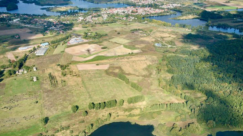 Luftaufnahme einer ländlichen Landschaft mit Feldern, Wäldern, Flüssen und verstreuten Gebäuden - ideal für Glamping oder als Ferienimmobilie - sowie einer entfernten Stadt unter einem teilweise bewölkten Himmel. 0121Management - der Campingplatz und Ferienimmobilien Makler