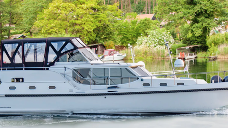 Eine weiße Motoryacht mit schwarzem Verdeck fährt auf einem ruhigen Fluss, vorbei an üppig grünen Bäumen und kleinen Häusern - die ideale Kulisse für ein Glamping oder die Erkundung Ihres Traum-Ferienobjekts entlang des Ufers. 0121Management - der Campingplatz und Ferienimmobilien Makler